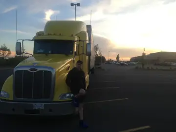Młody mężczyzna opiera się o żółtą ciężarówkę. W tle zachód słońca i parking pełen aut. To obrazek z życia polskie truckerki.